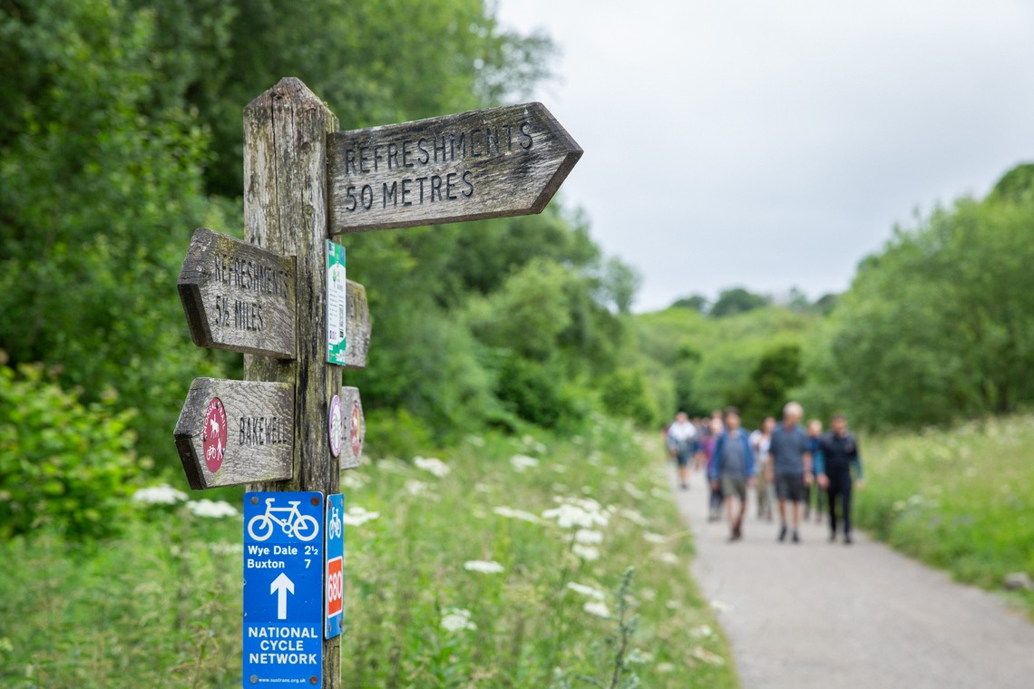 A wooden signpost displays directions, including "Refreshments 50 Metres," while a group of people walks along a winding path surrounded by lush greenery and wildflowers.