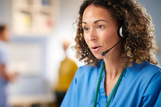 A woman in blue scrubs is speaking into a headset, engaged in a conversation. In the background, blurred figures in medical attire suggest a healthcare environment.
