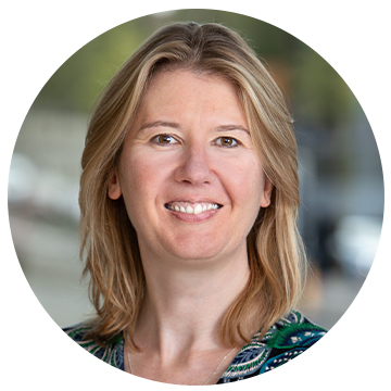 A smiling woman with medium-length blonde hair is positioned in a circular frame. She is facing the camera, set against a blurred outdoor background that includes hints of a cityscape.