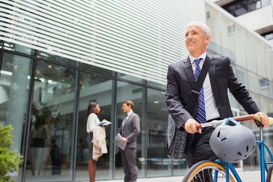 A man in a suit smiles while walking a bicycle, equipped with a helmet, through a modern outdoor area where two people converse, surrounded by glass buildings and greenery.