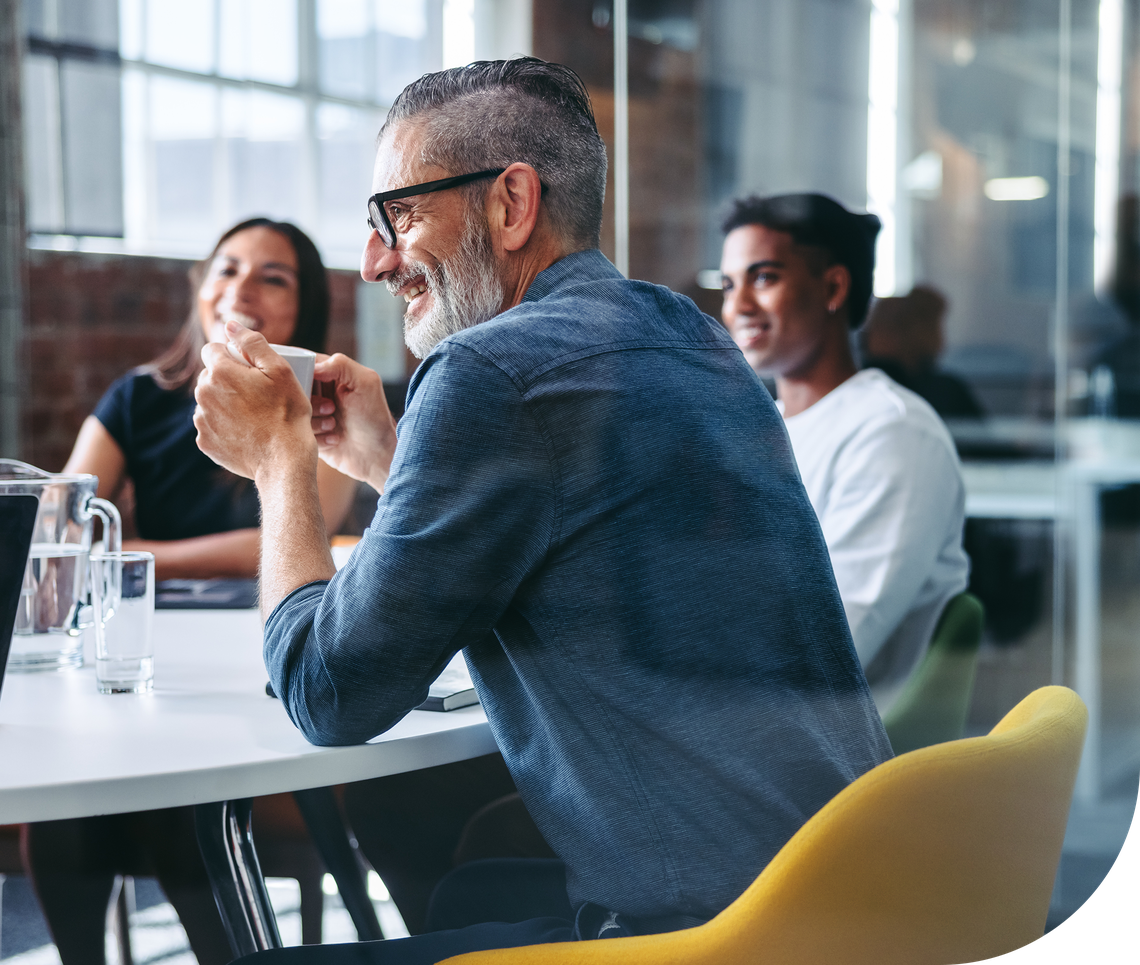 A man with a beard and glasses smiles while holding a cup, sitting at a round table with others in a bright, modern office setting featuring large windows and a casual atmosphere.