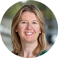 A woman with medium-length blonde hair smiles at the camera. She is dressed in a colorful patterned outfit, set against a blurred outdoor background featuring greenery and city elements.