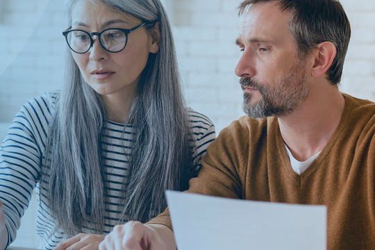 Two people, a woman with long hair and glasses and a man, are focused on a document while seated at a table in a bright, modern indoor space with light-colored walls.
