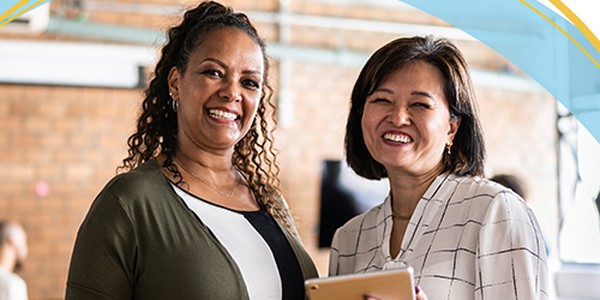 Two women smile while standing indoors, one holding a tablet. They appear to be engaged in a friendly conversation, with a brick wall and modern decor in the background.