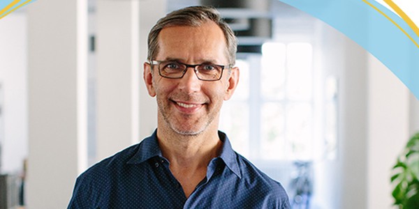 A smiling man with glasses stands in a bright, modern office space, framed by large windows and plants, giving a welcoming impression in casual attire.