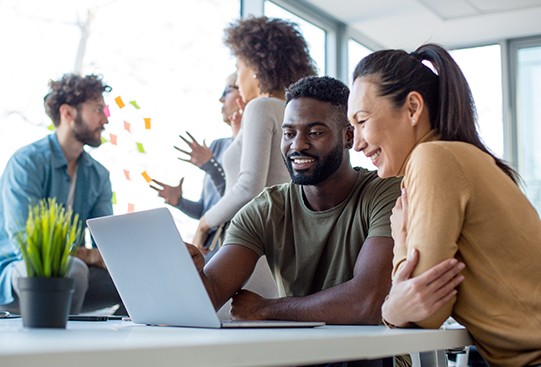 A man and woman share a laptop, smiling and engaged in conversation. In the background, four people engage in discussions, with colorful sticky notes on a wall, in a bright, modern office.