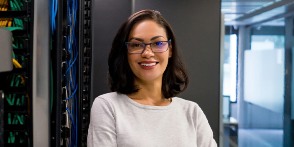 A woman with shoulder-length hair, wearing glasses and a gray sweater, smiles confidently while standing beside server racks in a modern, well-lit tech environment.
