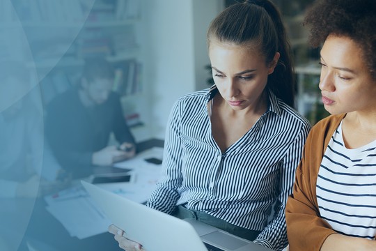 Two women, one in a striped shirt and the other in a brown sweater, are observing a document on a laptop, concentrated, in a modern workspace with blurred activity in the background.