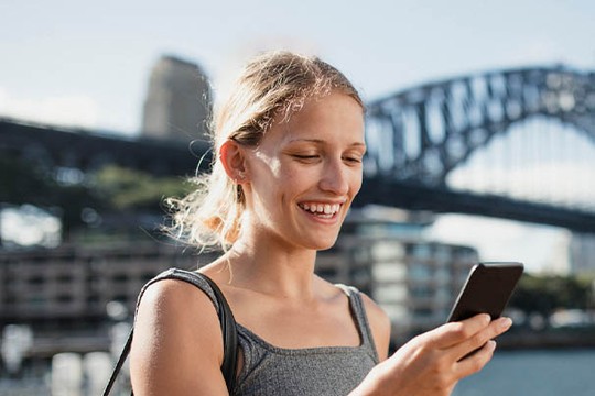 A young woman with shoulder-length hair smiles while looking at her smartphone. Behind her, a large bridge and urban buildings are visible under a clear blue sky.