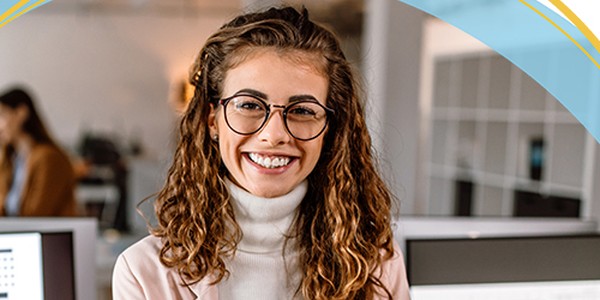 A woman with curly hair and glasses smiles warmly at the camera while standing in a modern office. Computer screens are visible in the background, indicating a professional environment.