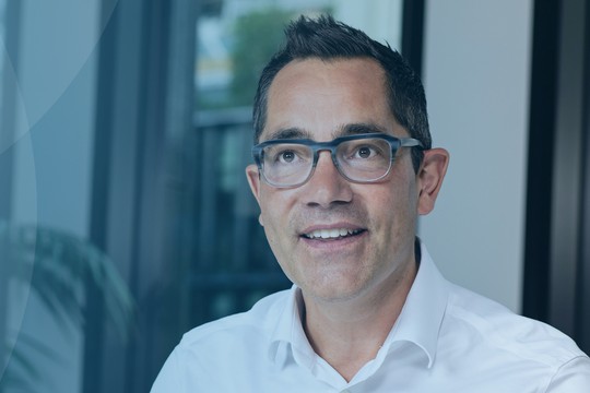 A man wearing glasses and a white shirt smiles while sitting in a well-lit indoor space. The background features blurred windows and greenery, creating a professional atmosphere.