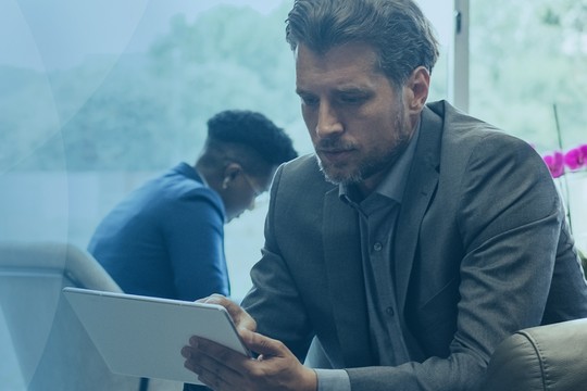 A man in a suit sits on a couch, intently using a tablet. In the background, two others are engaged in their own activities in a bright, modern space with greenery.