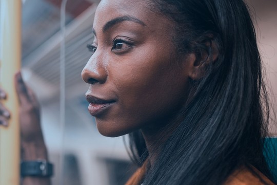 A woman is standing in a subway car, holding a pole, and gazing thoughtfully into the distance. The interior appears modern, with soft lighting and blurred figures in the background.
