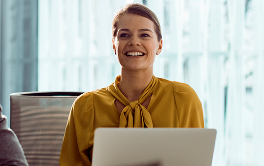 A woman in a yellow blouse smiles while seated at a conference table with two others. They are engaged in discussion in a bright, modern office environment with large windows.