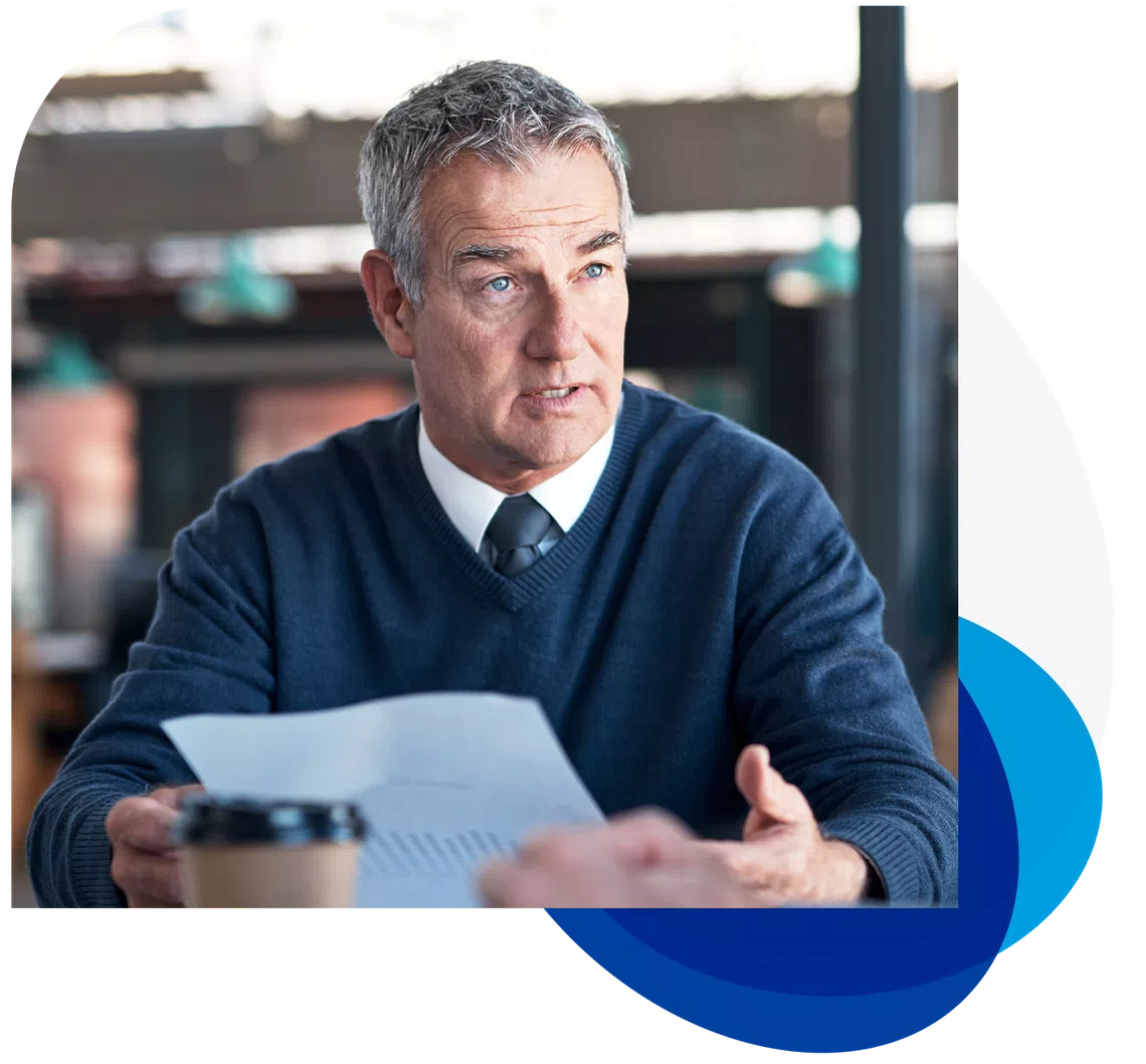A middle-aged man in a blue sweater and tie is holding a sheet of paper, speaking thoughtfully in a modern workspace with soft lighting and a coffee cup nearby.