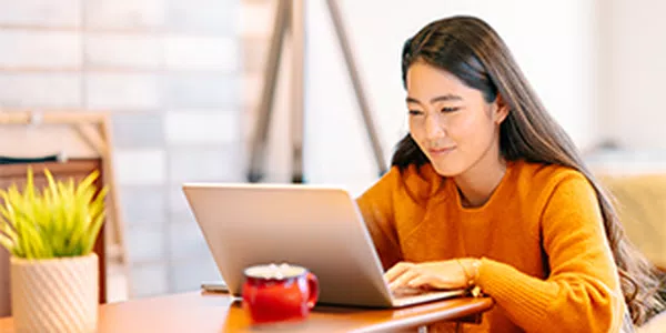 A woman sits at a wooden table, typing on a laptop with a focused expression. A small potted plant and a red mug are nearby, set in a bright, cozy indoor space.