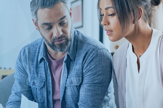 A man and woman sit closely on a sofa, reviewing documents. The room is bright and modern, featuring a plant and stylish decor in the background.