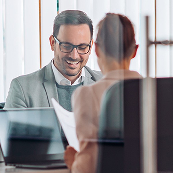A group of four professionals are engaged in a discussion around a table, smiling and looking at documents, in a modern office with large windows and a whiteboard.