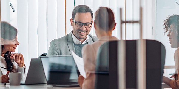 A group of four professionals are engaged in a discussion around a table, smiling and looking at documents, in a modern office with large windows and a whiteboard.