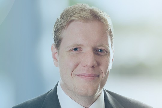 A man in a suit smiles confidently, facing the viewer against a soft blue gradient background. The context suggests a professional or business setting, highlighting a sense of approachability and professionalism.