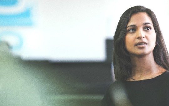 A woman speaks engagingly with a man in a professional setting, surrounded by office elements and blue graphics on the wall, conveying a collaborative atmosphere.