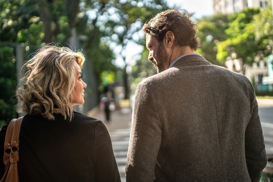 Two people walk closely together on a tree-lined street, sharing a moment. The woman, with blonde curly hair, smiles at the man beside her, who wears a gray coat.