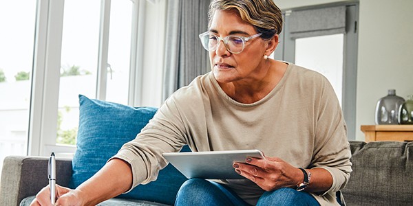 A woman sits on a couch, holding a tablet in one hand and writing with a pen in the other. Natural light streams through large windows, illuminating the modern, cozy space.