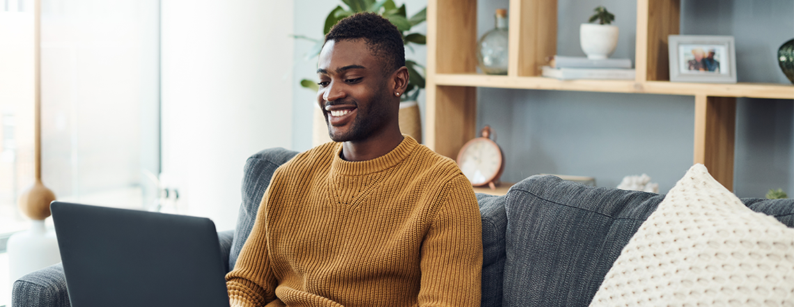 A man in a cozy, mustard-colored sweater is smiling while using a laptop. He sits on a gray sofa in a bright, modern living room with plants and decorative shelves.