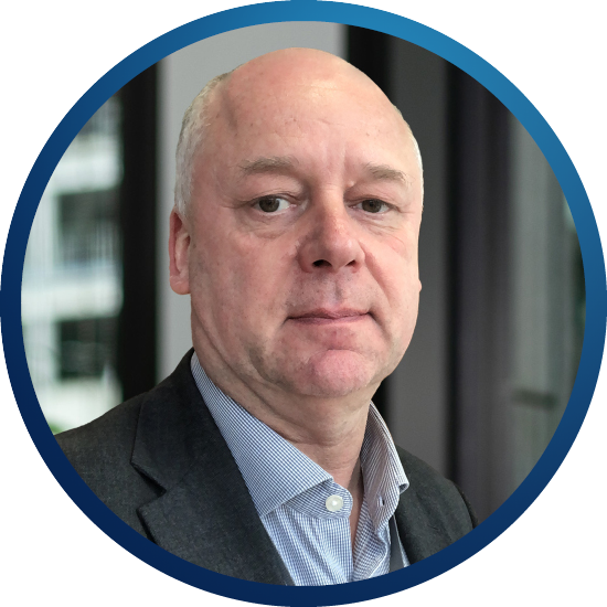 A man with short hair and a neutral expression gazes directly at the camera. He wears a dark suit and light blue shirt, set against a blurred indoor background.