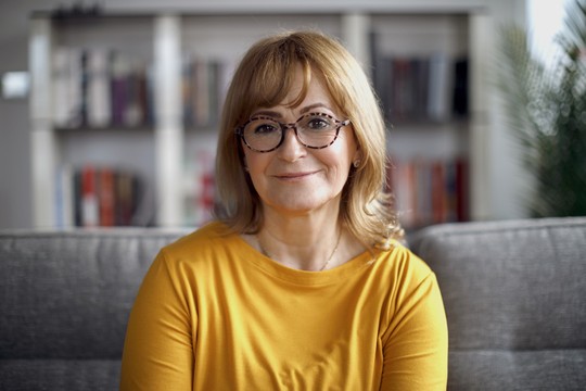 A woman with blonde hair, wearing round glasses and a yellow top, smiles while seated on a gray couch, surrounded by shelves filled with books in a bright room.