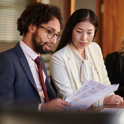 A man and a woman review documents in an office setting, while a third woman works on a tablet. The background features wooden panels and a coffee cup on the table.