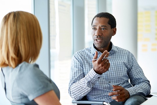 A man engaged in conversation holds a pen while expressing ideas to a seated woman. They are in a modern office space with large windows and visible sticky notes on the wall.