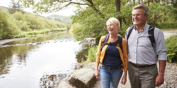 A man and woman walk along a riverbank, smiling and talking. They wear backpacks, surrounded by lush greenery and trees, with calm water reflecting the landscape.