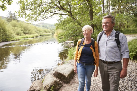 A man and woman walk along a riverbank, smiling and talking. They wear backpacks, surrounded by lush greenery and trees, with calm water reflecting the landscape.