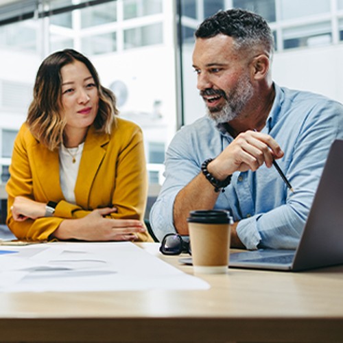 A man and a woman engage in discussion at a table cluttered with papers, while a laptop and a coffee cup sit nearby. Large windows allow natural light to fill the modern workspace.
