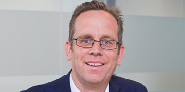 A man with glasses smiles, wearing a suit against a softly blurred background of light-colored walls, suggesting a professional or office setting.