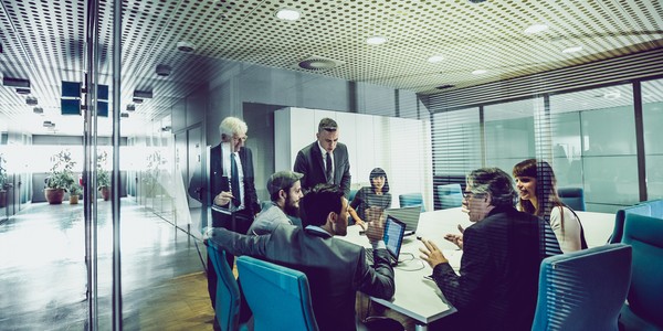 A group of professionals engages in a discussion around a conference table, with multiple laptops visible. The setting is an office space with glass walls and modern furnishings.
