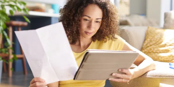 A woman with curly hair sits on the floor, holding a document in one hand and a tablet in the other, absorbed in reading while surrounded by a cozy living space.