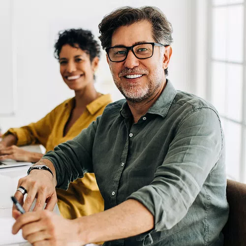 A middle-aged man with glasses smiles while holding a pen. A woman with curly hair, seated behind him, appears cheerful. They are in a bright office setting.
