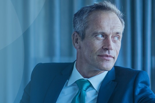 A middle-aged man in a suit gazes thoughtfully to the side. He sits in a modern office, characterized by soft blue tones and sleek furnishings, with light filtering through glass panels.
