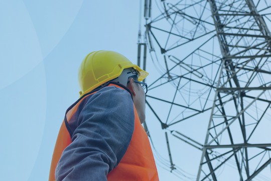 A worker in safety gear gazes upward at a tall power transmission tower against a clear blue sky, reflecting focus on electrical infrastructure maintenance or inspection.