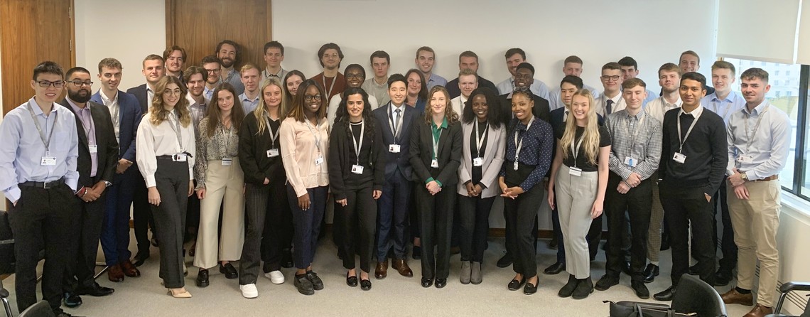 A diverse group of over 40 individuals stands together in an office setting, all smiling toward the camera, wearing name tags and professional attire, with wooden walls and windows in the background.