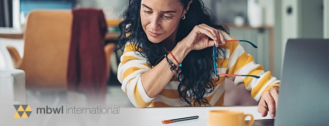 A woman in a striped sweater adjusts her glasses while focused on writing or drawing at a desk, surrounded by everyday objects like pens and a mug in a cozy, modern room.