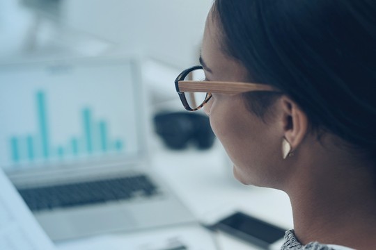 A woman with glasses examines printed documents while seated at a desk. A laptop displays a bar graph in the background, indicating a workspace focused on analysis.