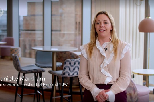 A woman stands and speaks confidently in a modern, well-lit office space with stylish furniture, including small tables and colorful chairs, visible in the background. 

**Text:**
Danielle Markham  
Principal and Head of LDI Research, BW