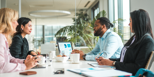 Five professionals engage in a discussion around a conference table, with laptops and coffee cups present. Large windows show a bright, modern office space filled with plants.