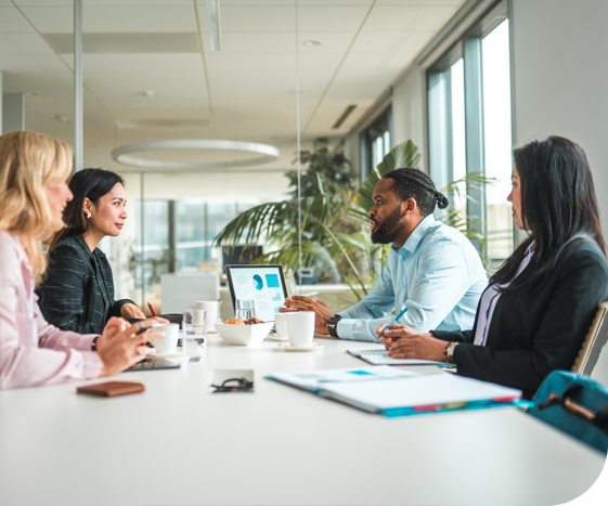 Five professionals engage in a discussion around a conference table, with laptops and coffee cups present. Large windows show a bright, modern office space filled with plants.