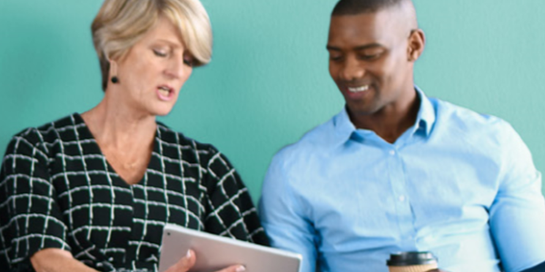 A woman and a man are seated together, reviewing content on a tablet. The woman appears engaged while the man smiles, holding a coffee cup, against a light green wall backdrop.