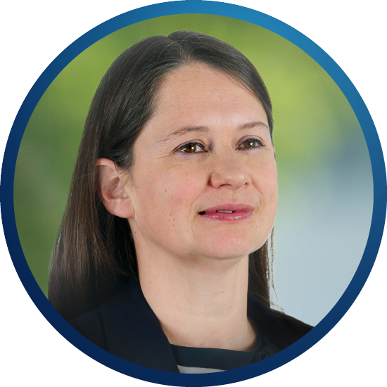 A woman with long, dark hair smiles gently. She is wearing a black jacket over a striped shirt, set against a blurred, greenish background that suggests an outdoor setting.