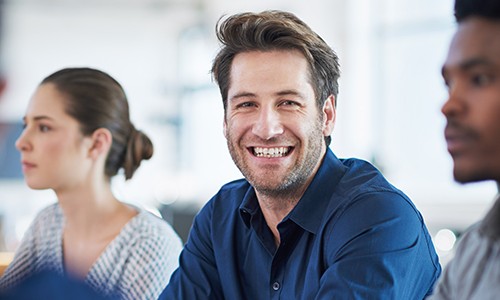 A smiling man in a blue shirt engages with others at a table. A woman in a patterned top and a man nearby listen attentively in a well-lit, modern workspace.